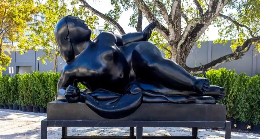 Reclining bronze sculpture displayed outdoors among trees and landscaping.